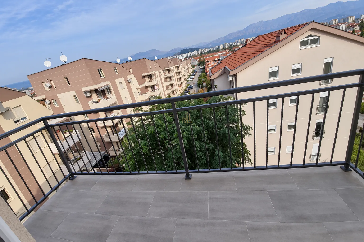 Pogled sa popločanog balkona sa crnom metalnom ogradom na okolne zgrade i planine.