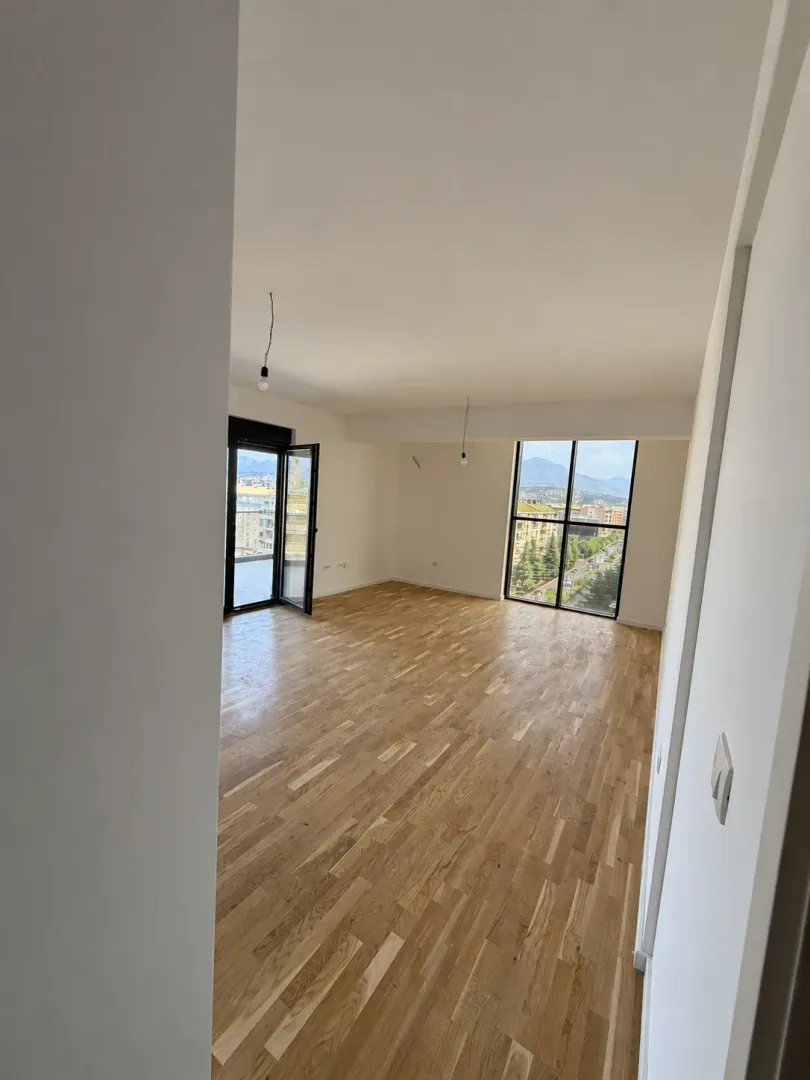 Prazna dnevna soba sa parketom i velikim prozorima sa pogledom na grad.