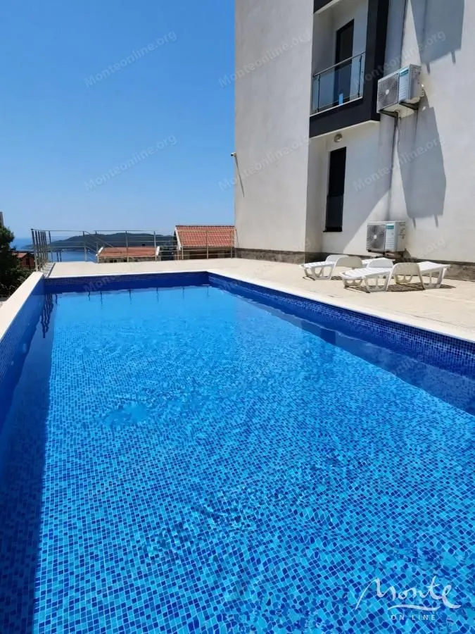 Spoljni bazen sa popločanim platoom ispred stambene zgrade i pogledom na more.