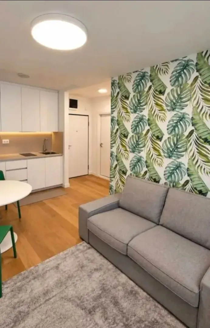 Living room with a sofa and botanical-themed wallpaper, alongside a kitchenette.
