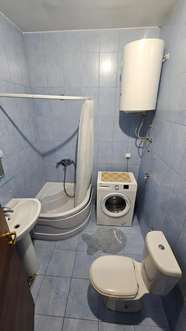 Manje kupatilo sa plavim pločicama, tuš kabinom, umivaonikom, veš mašinom i toaletom.