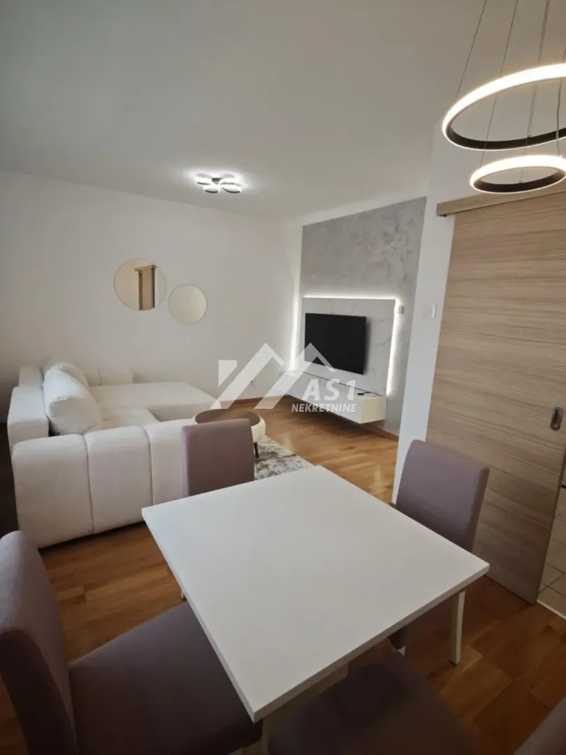 Dnevna soba sa ugaonom garniturom, trpezarijskim stolom i televizorom na zidu.