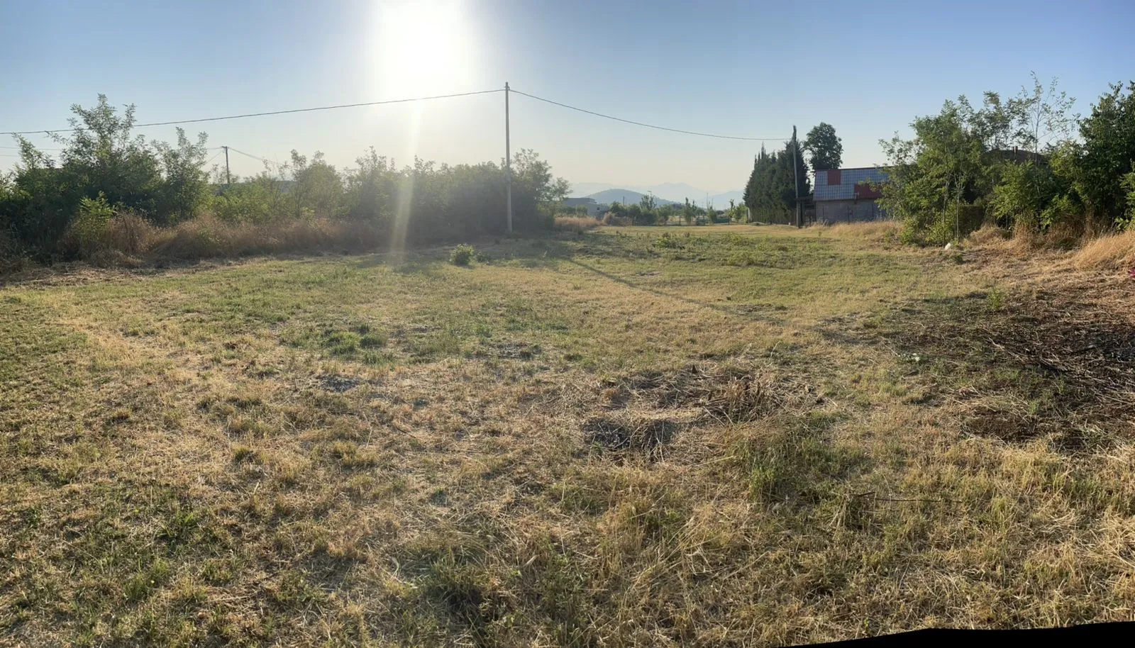 Pogled na prostrani ravni plac obrasao travom i niskim rastinjem sa drvećem u pozadini.