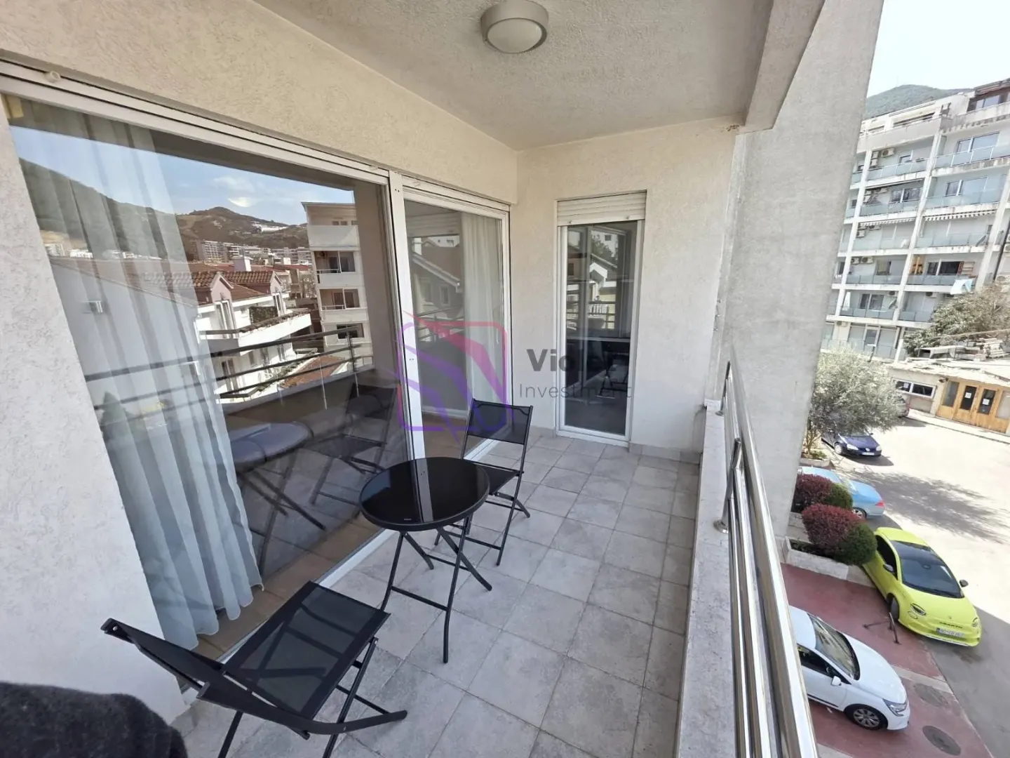 Covered balcony with seating furniture and a view of surrounding buildings and parking.