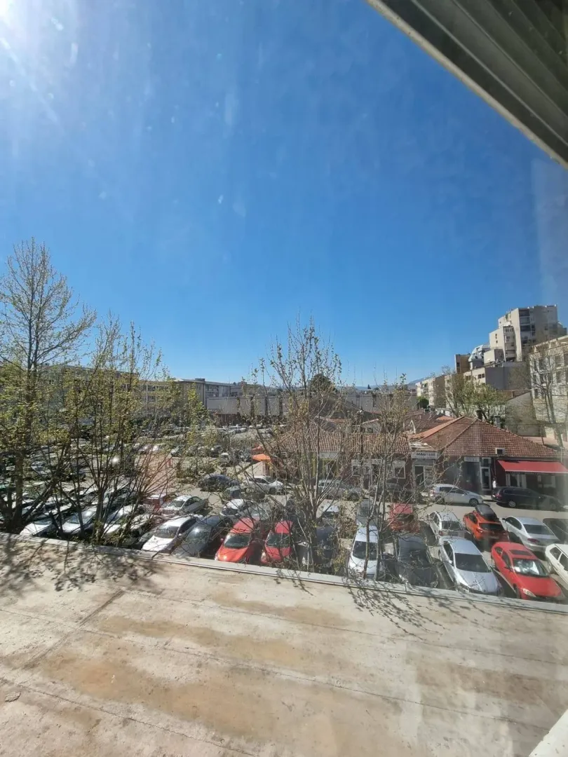 Pogled sa prozora na parking i okolne zgrade pod vedrim nebom.
