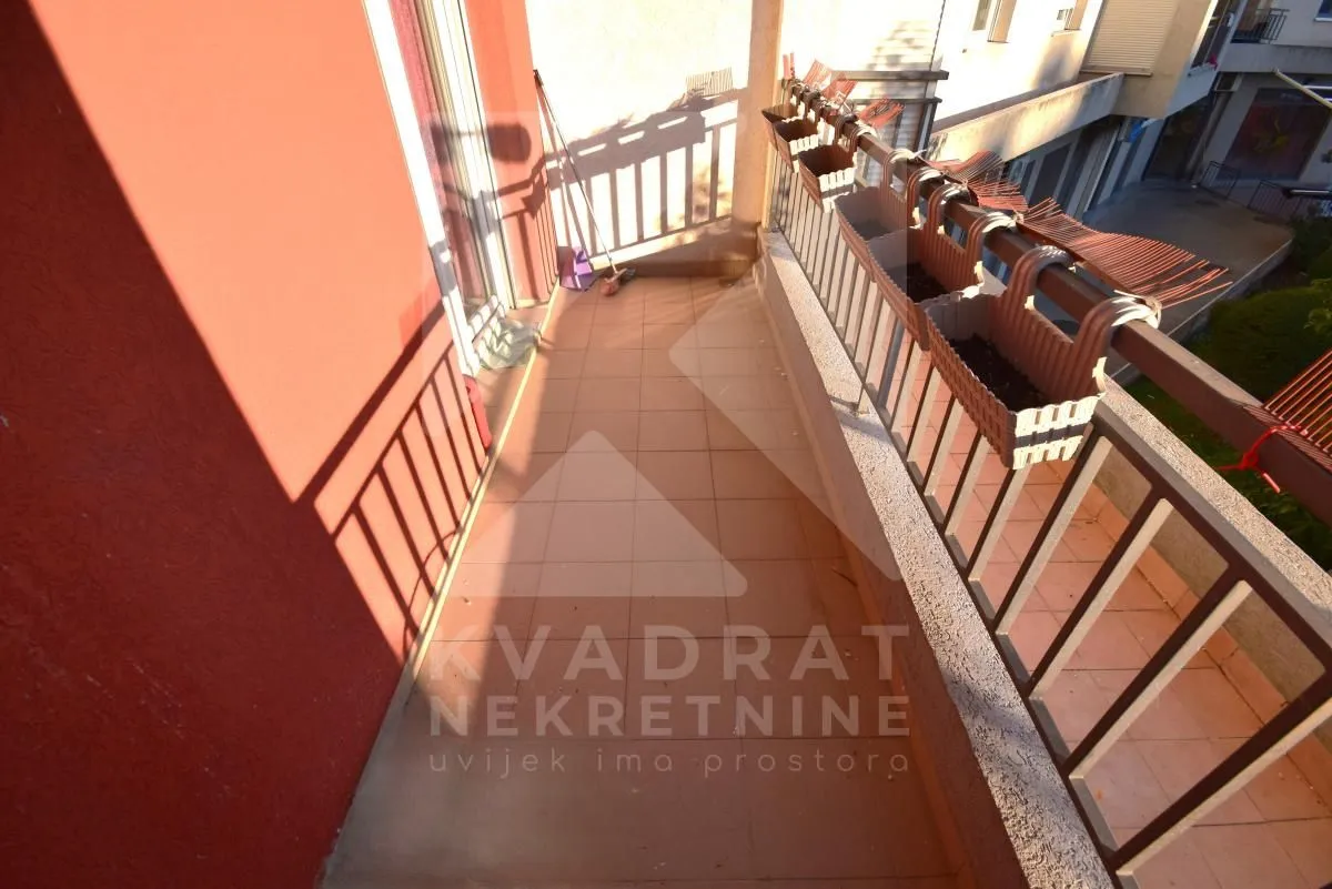 Dugi popločani balkon sa metalnom ogradom i žardinjerama za cvijeće.