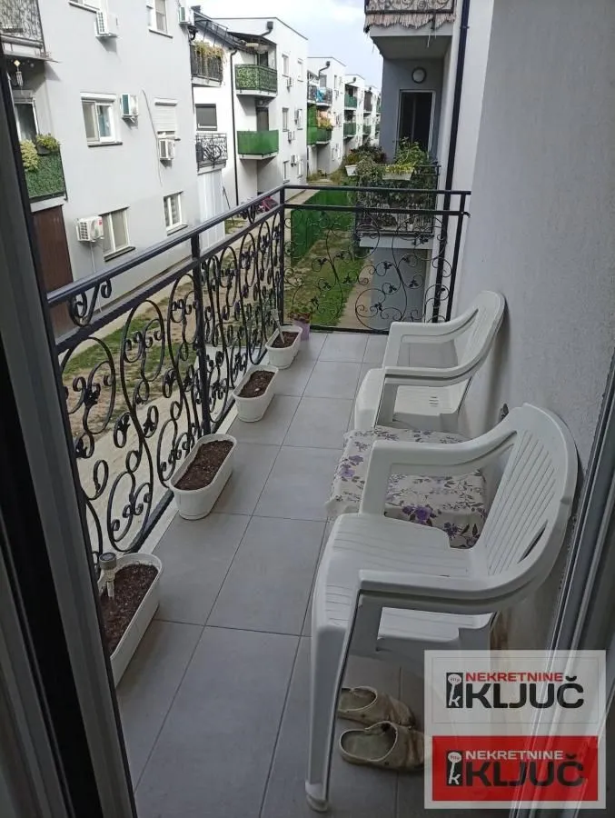 Uski popločani balkon sa ukrasnom ogradom, plastičnim stolicama i saksijama sa biljkama.