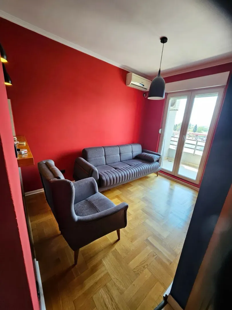 Dnevna soba sa crvenim zidom, garniturom i izlazom na balkon.