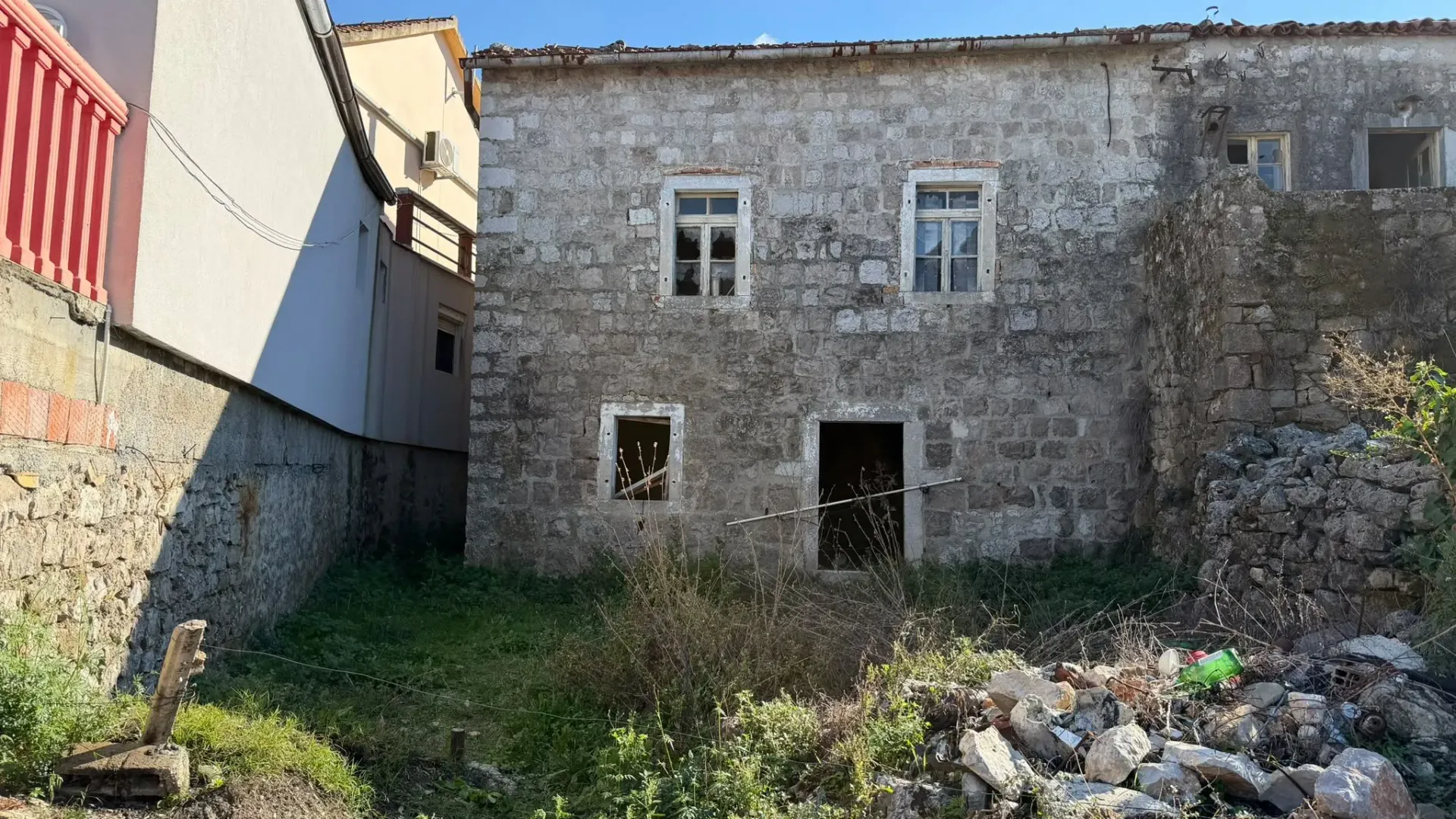 Fasada stare kamene kuće sa dvorištem koje je zaraslo u korov.