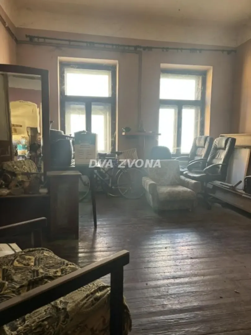 Stariji dnevni boravak sa drvenim podom, starim nameštajem i dva prozora.