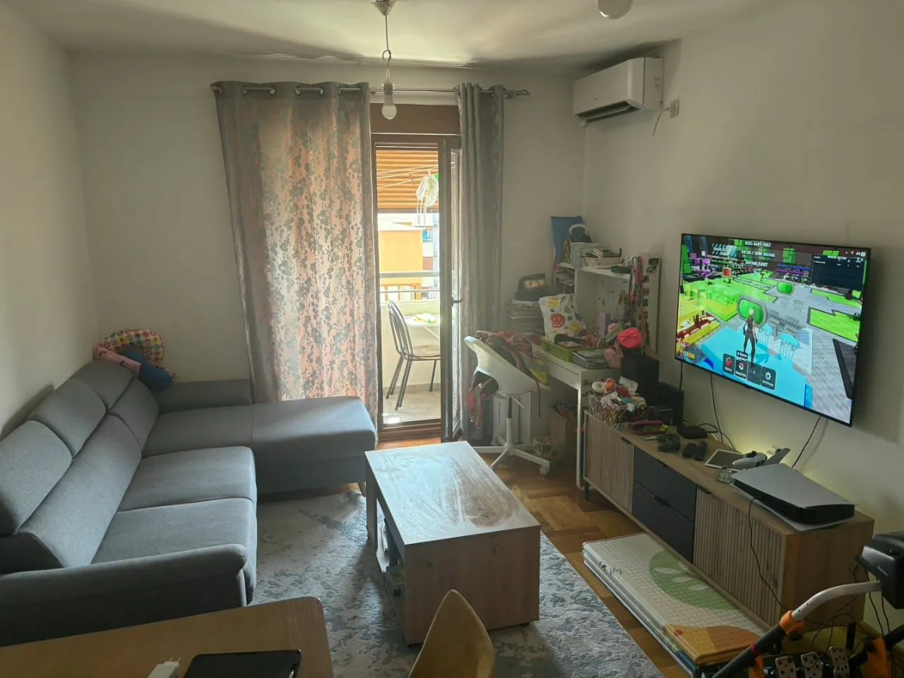Dnevna soba sa ugaonom garniturom, televizorom i izlazom na balkon.