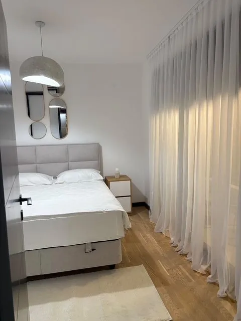 Svijetla spavaća soba sa bračnim krevetom i velikim prozorima prekrivenim zavjesama.