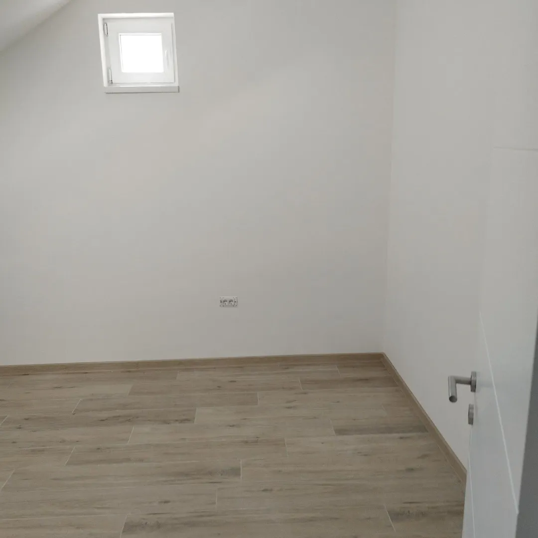 Prazna soba sa belim zidovima, drvenim podom i malim krovnim prozorom.