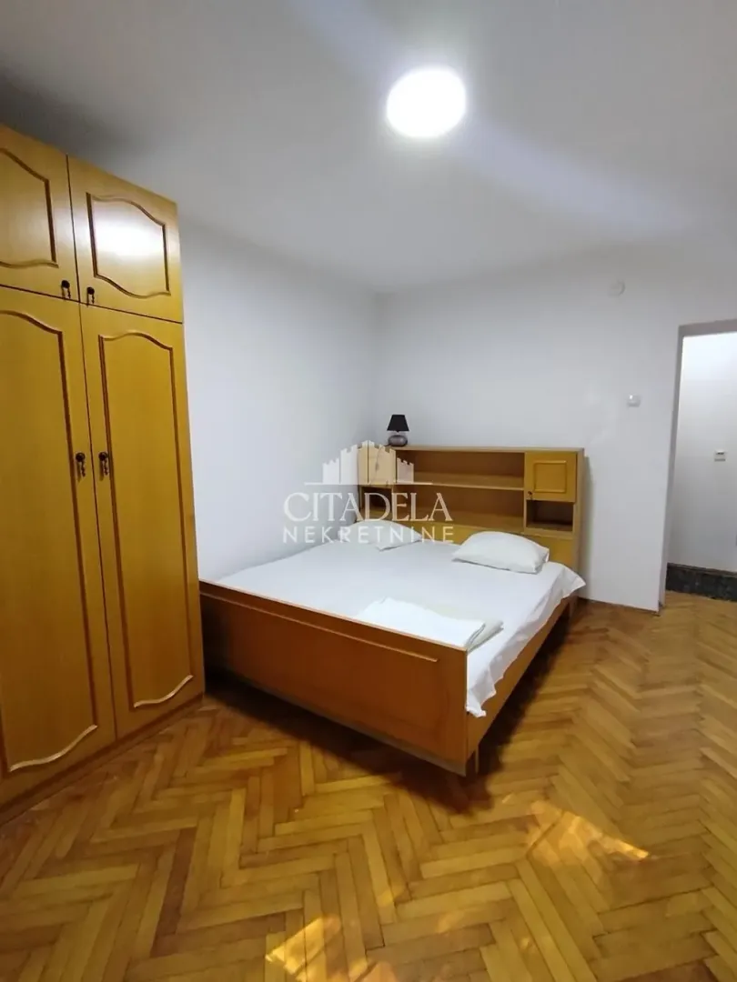 Nameštena spavaća soba sa bračnim krevetom, drvenim ormarom i parketom na podu.