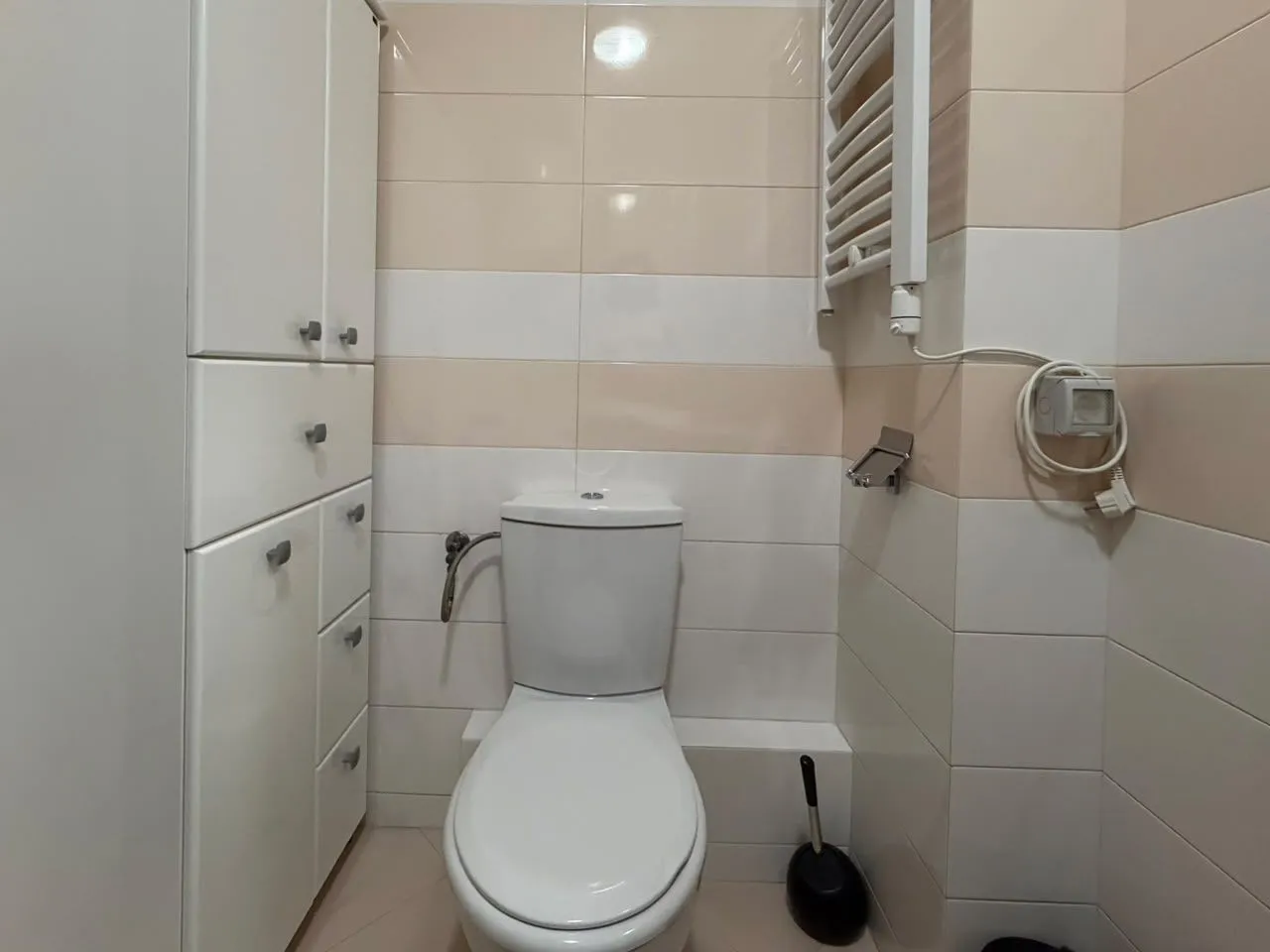 Manje kupatilo sa bijelim ormarićem, WC šoljom i radijatorom na zidu.