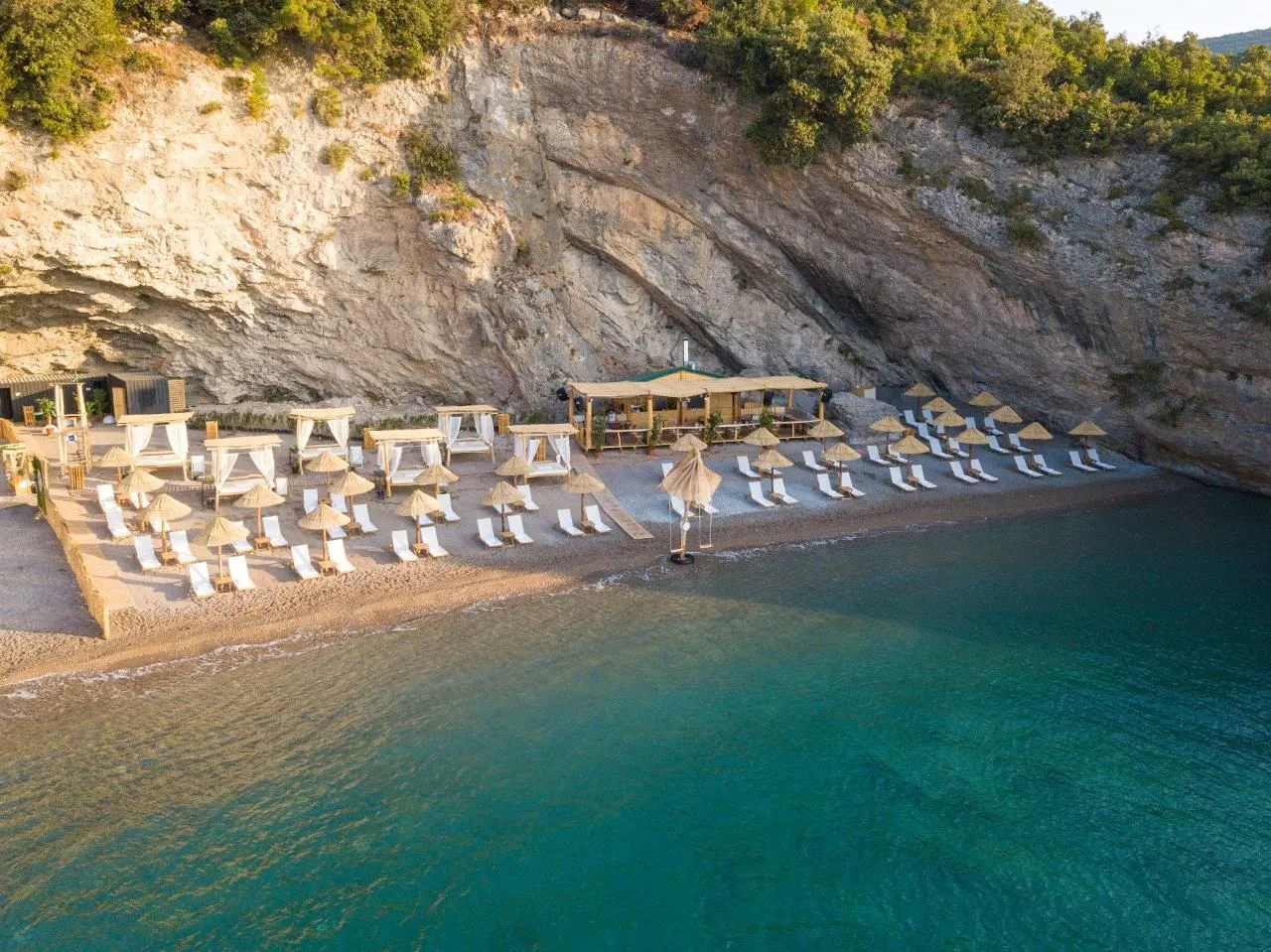 Pogled iz vazduha na uređenu plažu sa ležaljkama i suncobranima ispod stijene.