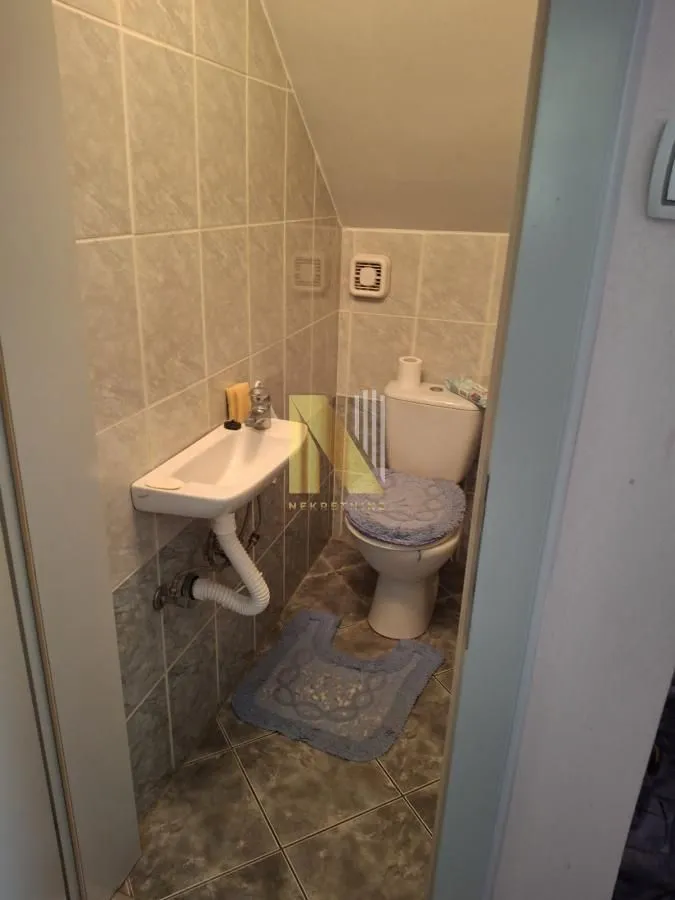 Manje kupatilo sa lavaboom, wc šoljom i zidnim pločicama pod kosinom krova.