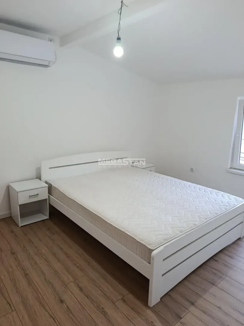 Svetla spavaća soba sa belim krevetom, noćnim stočićem i klima uređajem.