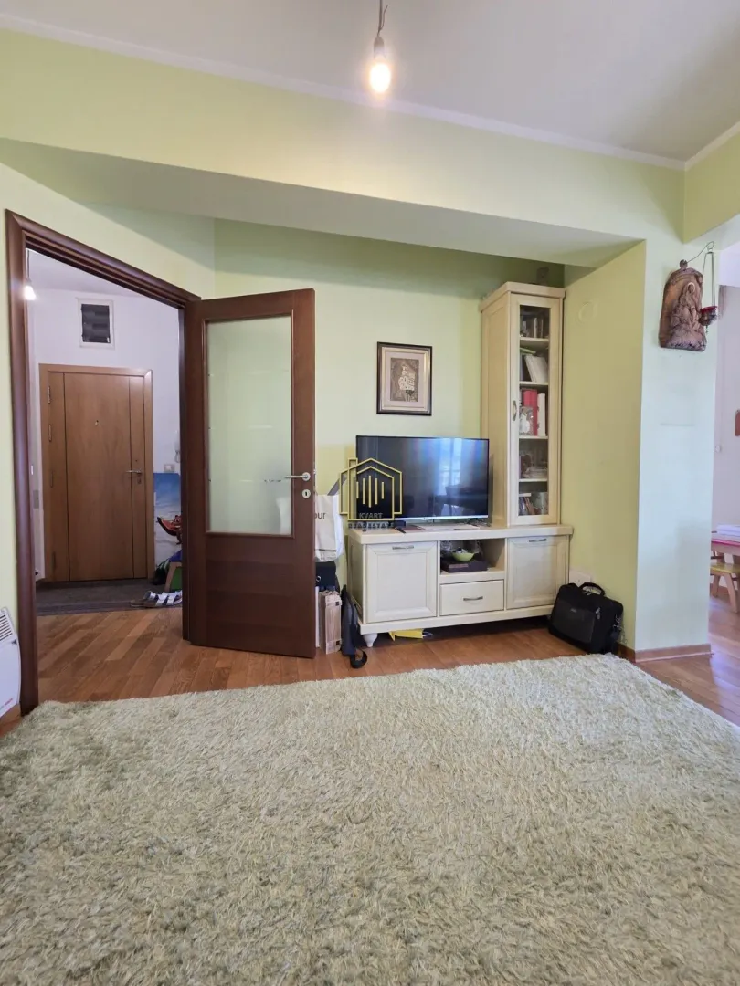 Dnevna soba sa TV komodom i tepihom, sa pogledom na hodnik.