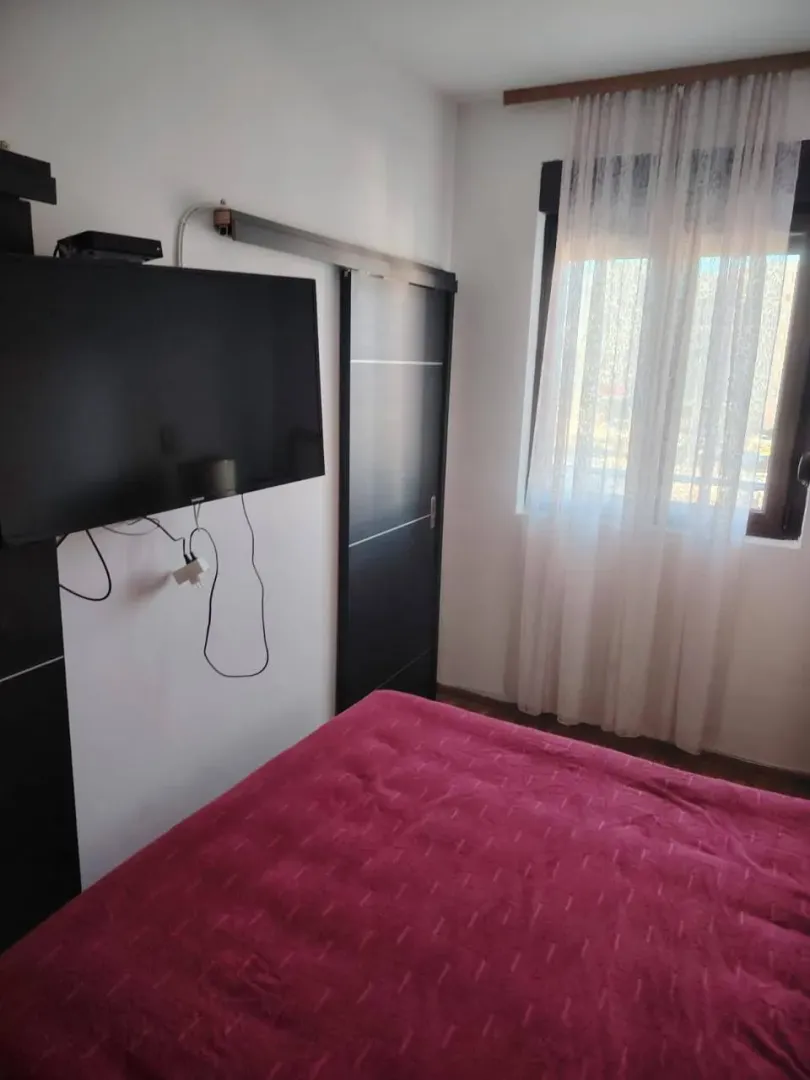 Spavaća soba sa bračnim krevetom, televizorom na zidu i prozorom sa zavjesama.