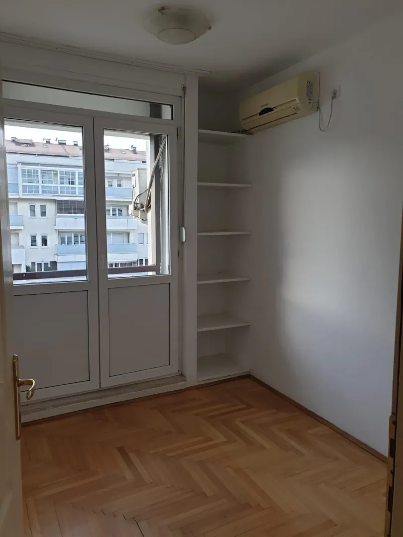 Empty room with parquet flooring, built-in shelves, and an air conditioning unit next to balcony doors.