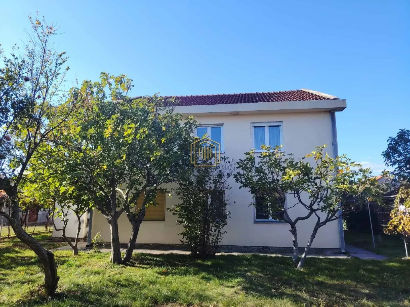 Pogled na dvorište i fasadu kuće sa zelenilom i stablima.