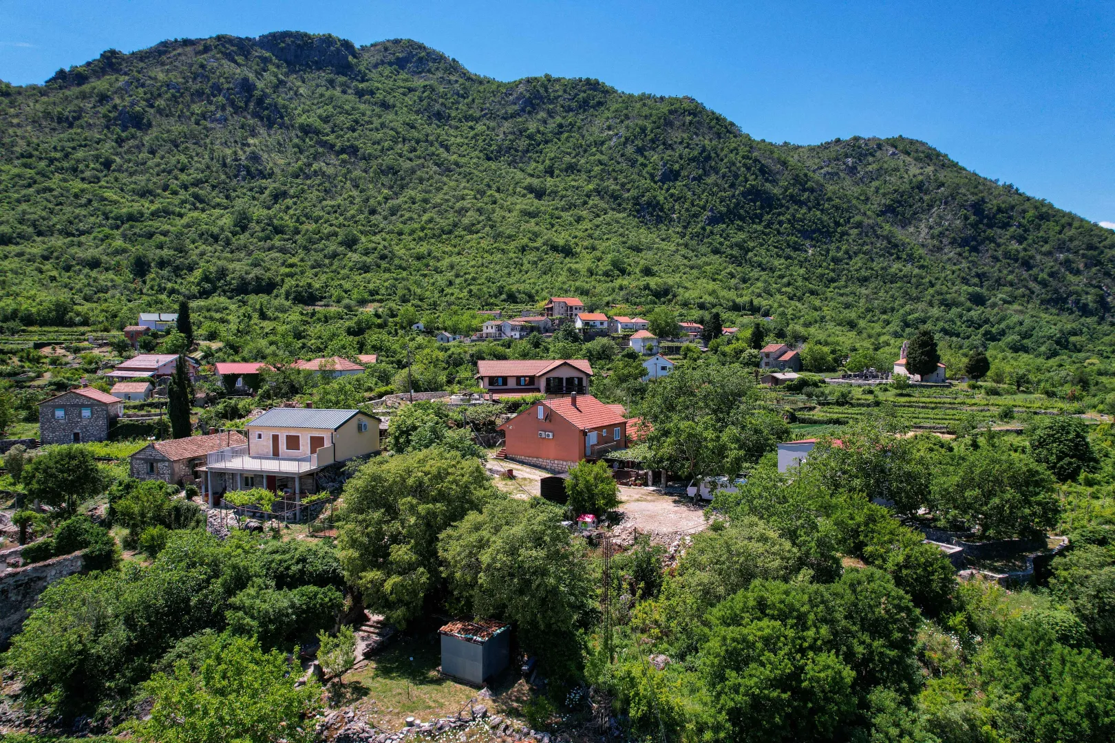 Pogled iz vazduha na seosko naselje pod brdom obraslim šumom.