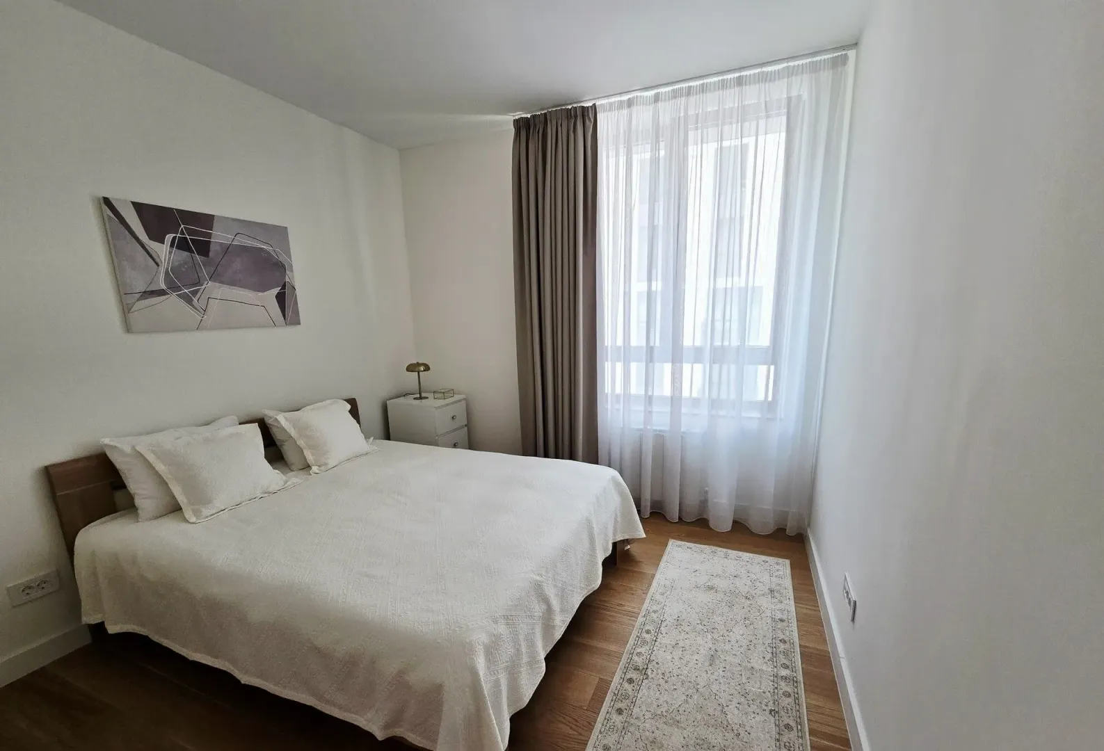 Svetla spavaća soba sa bračnim krevetom, noćnim stočićem i prozorom prekrivenim zavesama.