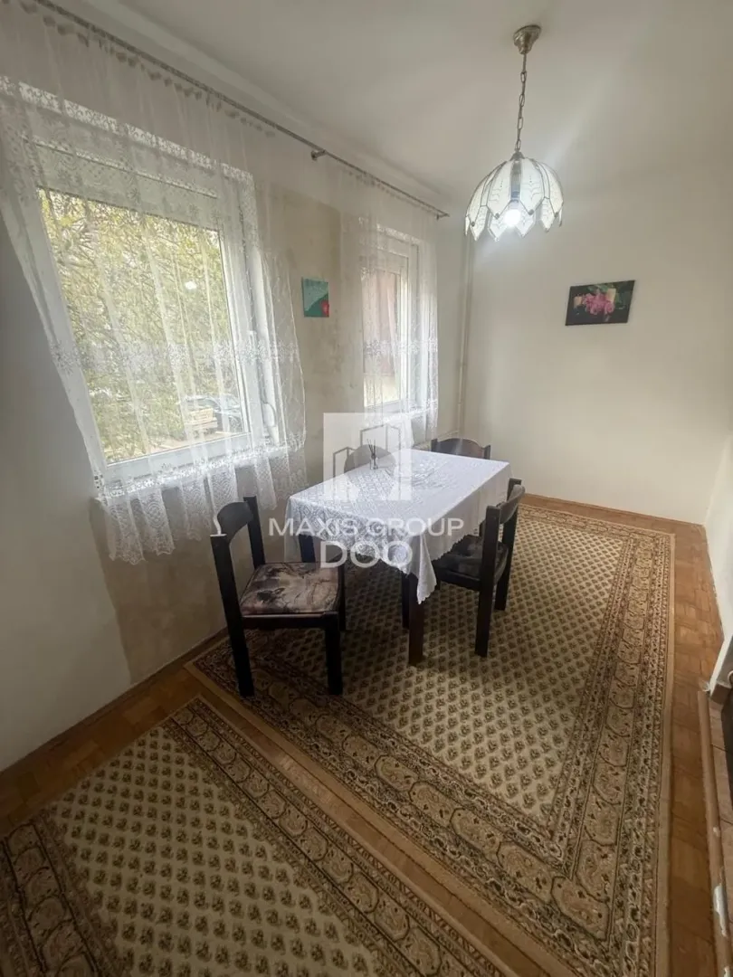 Trpezarija sa stolom, stolicama i dva prozora sa belim zavesama.