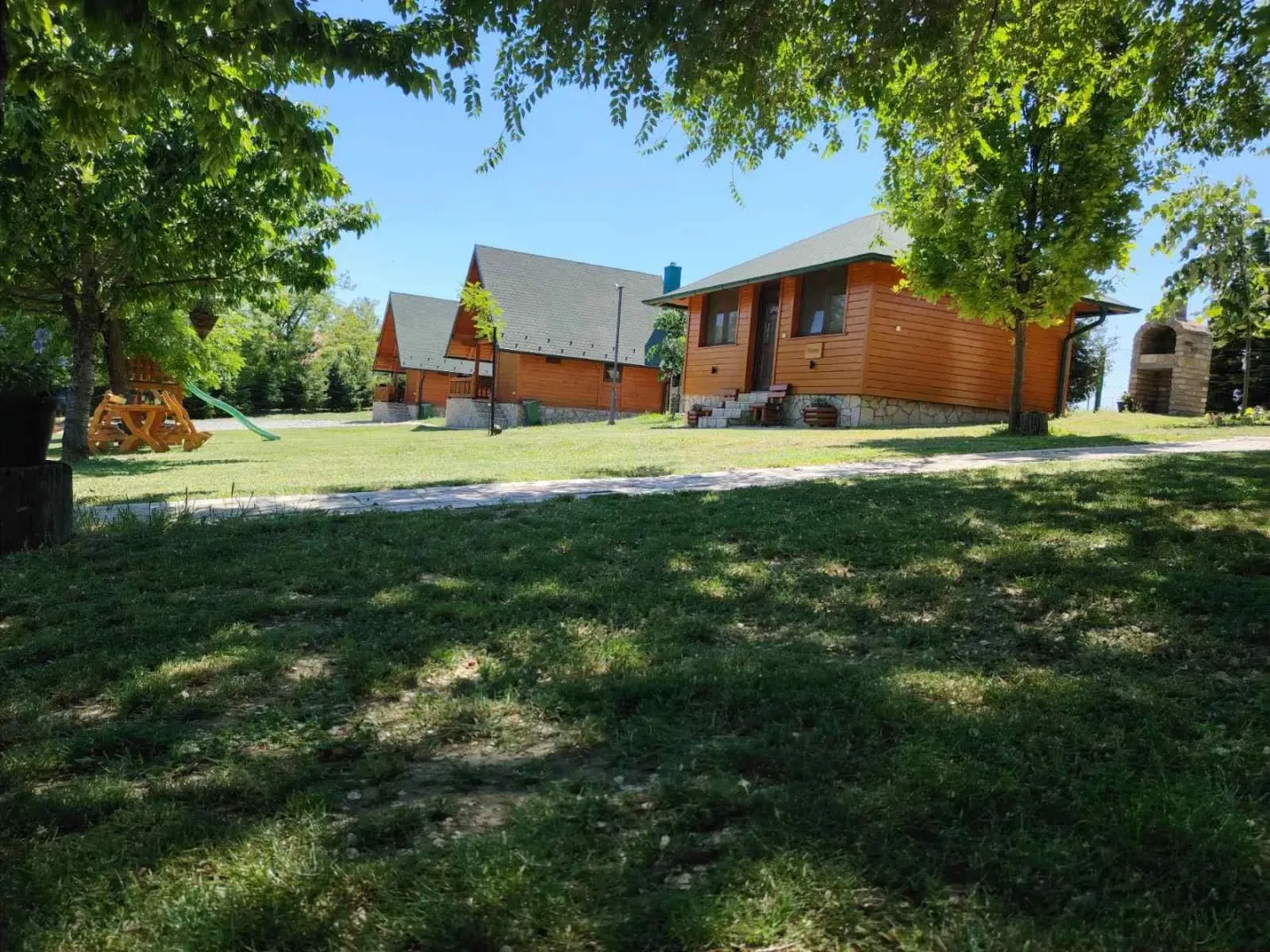 Pogled na drvene kuće u uređenom dvorištu sa travnjakom i drvećem.