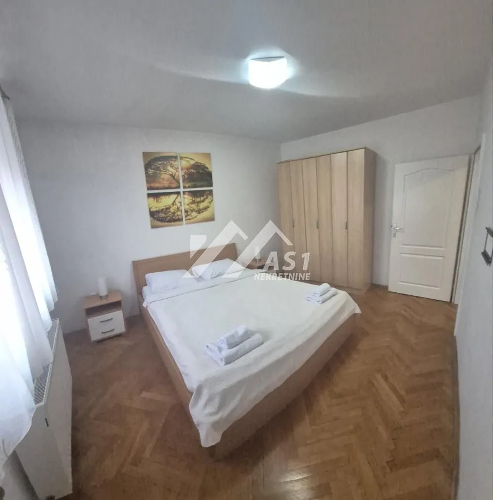 Nameštena spavaća soba sa bračnim krevetom, ormanom i drvenim podom.
