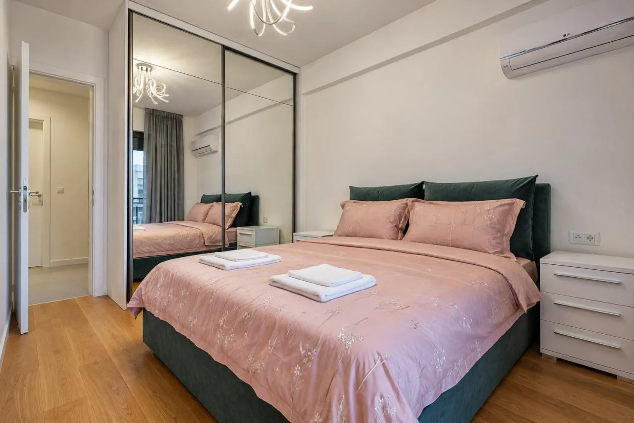 Furnished bedroom with a double bed, large mirrored wardrobe, and air conditioning unit.
