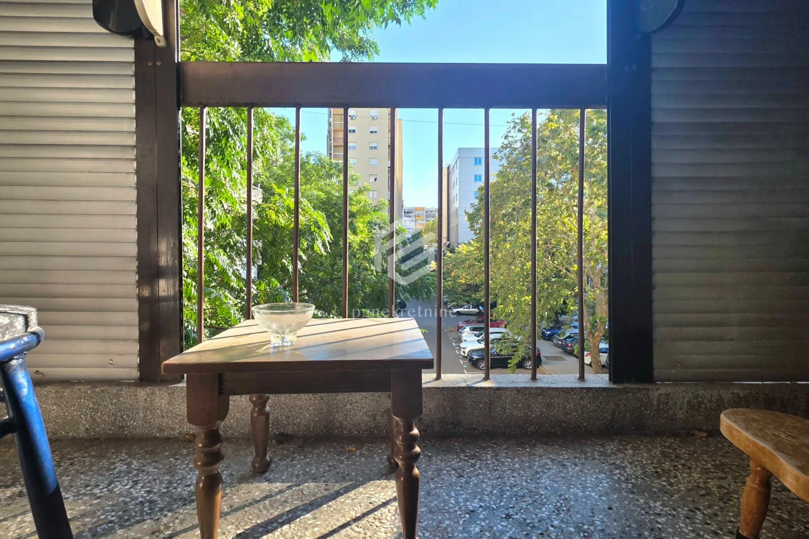 Covered balcony with a wooden table and a view of the street and greenery.