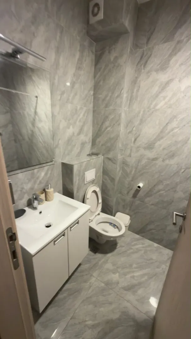 Manje kupatilo obloženo sivim pločicama sa umivaonikom i wc šoljom.