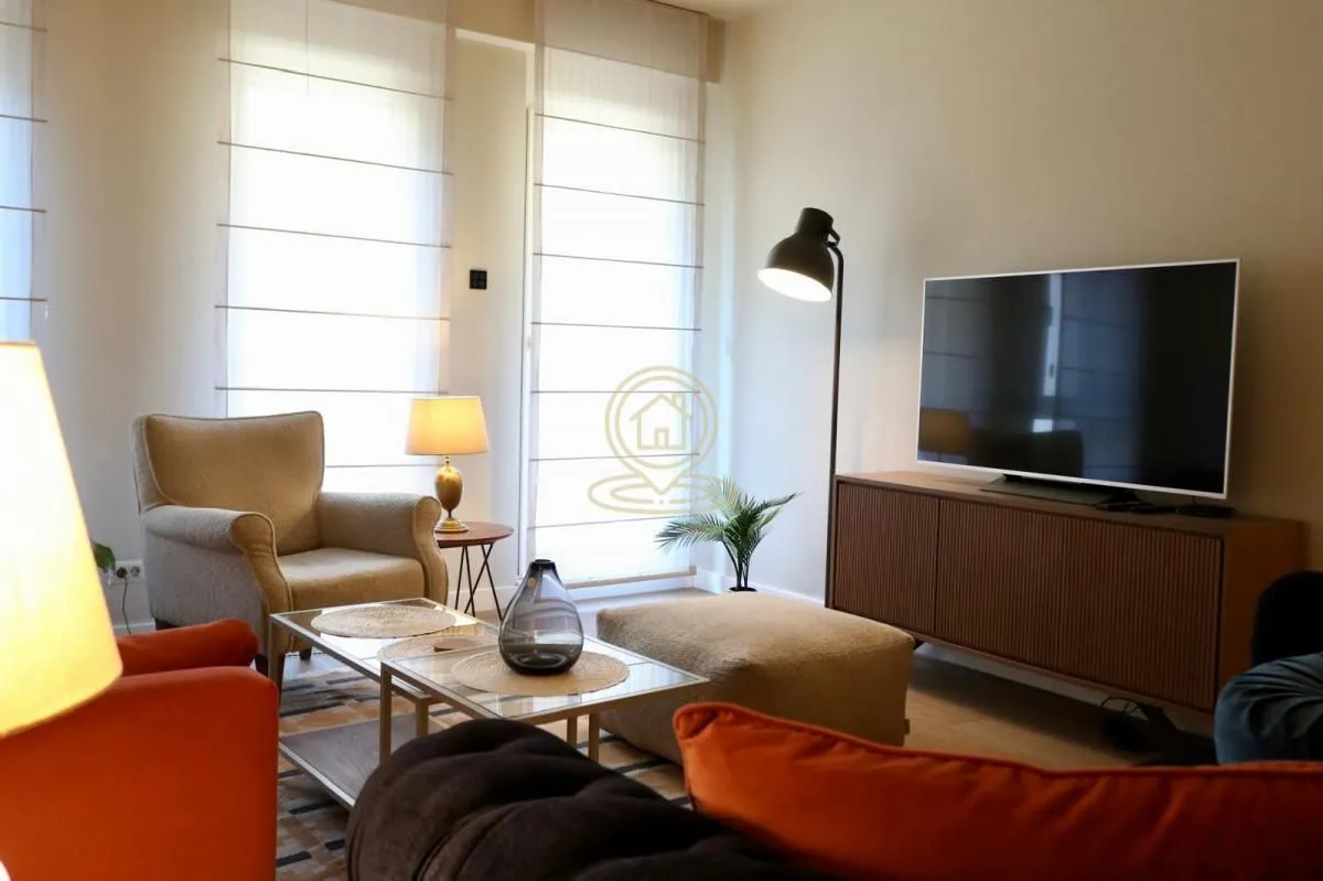 Udobna dnevna soba sa garniturom, foteljom, TV-om i velikim prozorima.