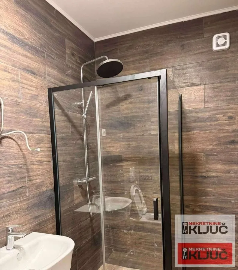 Kupatilo sa tuš kabinom i umivaonikom, obloženo keramičkim pločicama u dezenu drveta.