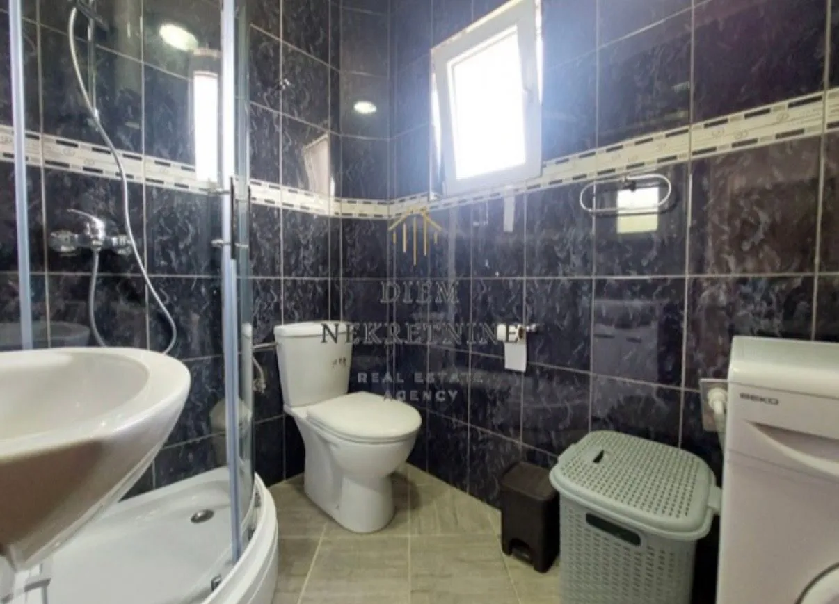 Kupatilo sa tuš kabinom, WC šoljom, umivaonikom i pločicama tamne boje.