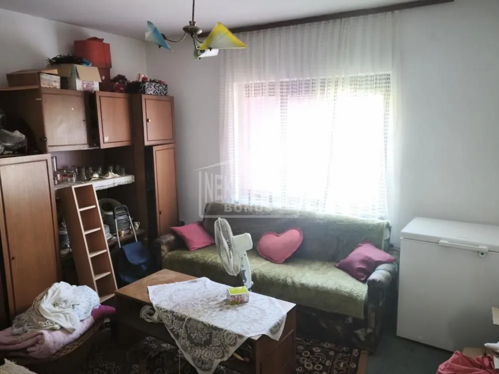 Dnevna soba sa garniturom za sedenje, drvenim nameštajem i prozorom sa belom zavesom.