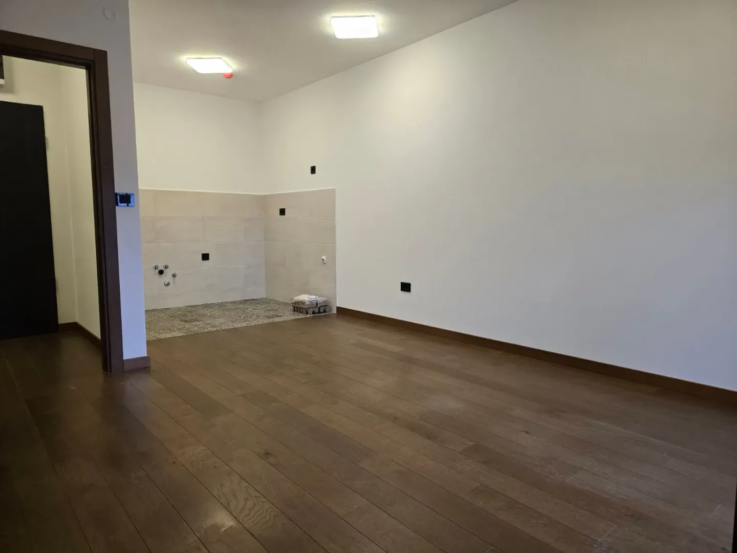 Empty room with kitchen connections and hardwood flooring.