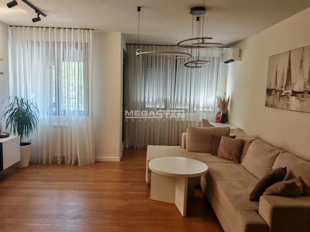 Dnevna soba sa ugaonom garniturom, stočićem i velikim prozorima sa belim zavesama.