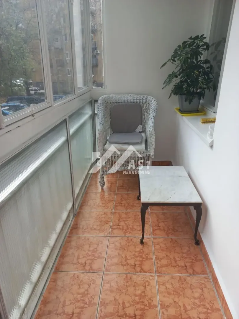 Zastakljeni balkon sa foteljom, stočićem i saksijom sa cvećem.