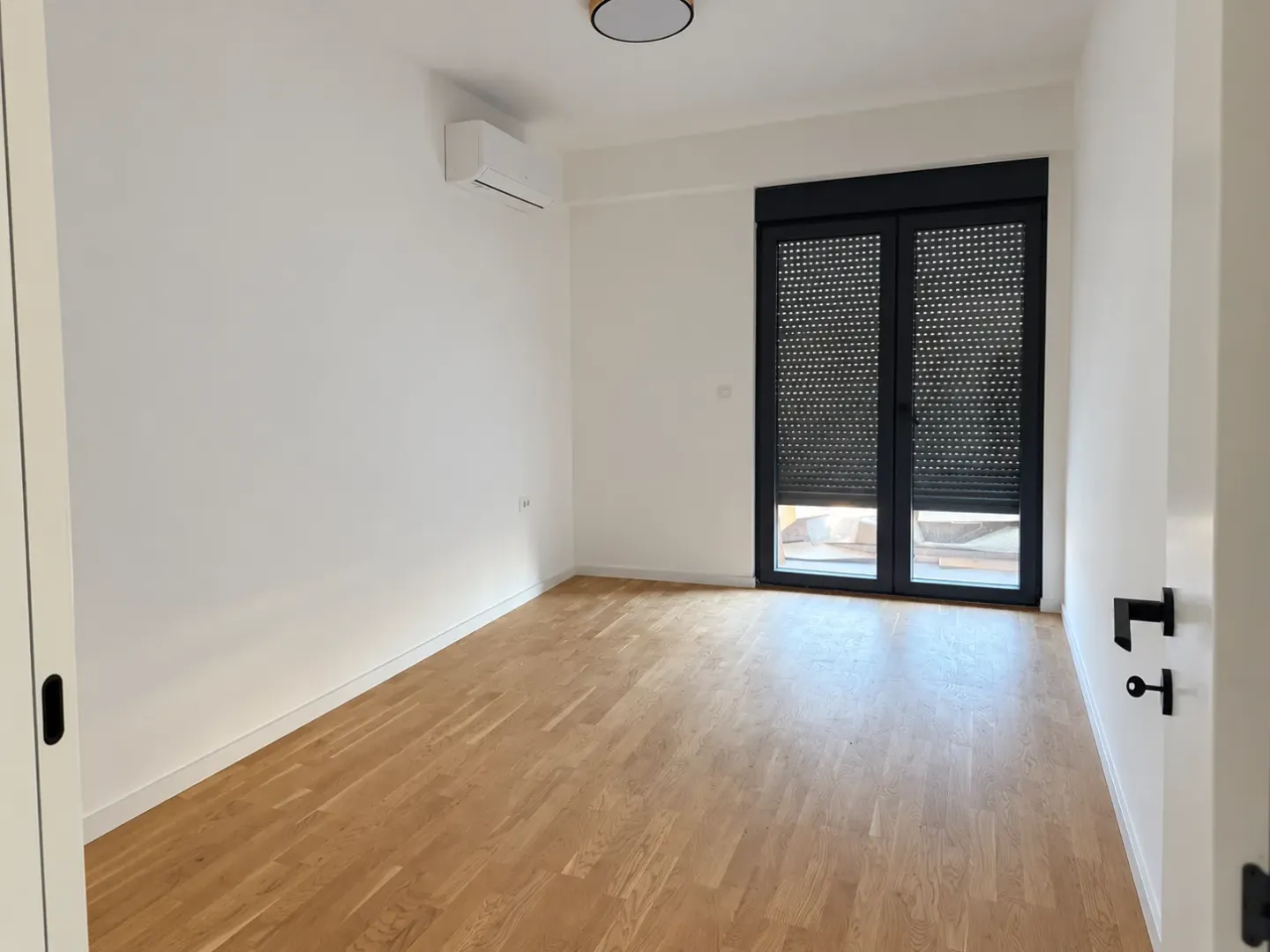 Prazna soba sa parketom, klima uređajem i balkonskim vratima sa roletnama.
