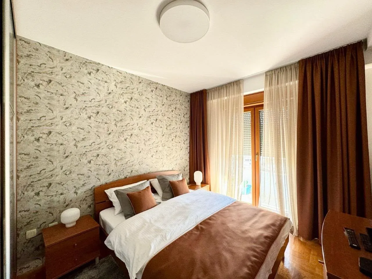 Furnished bedroom with a double bed, nightstands, and a large window with curtains.