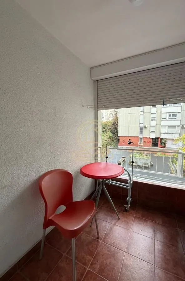 Manji zatvoreni balkon sa stočićem i crvenom stolicom i pogledom na zgrade.