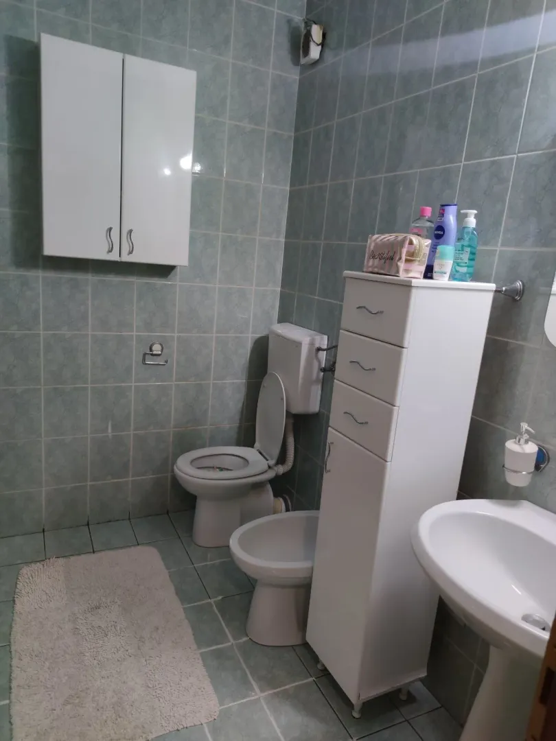 Kupatilo sa zelenim pločicama, toaletom, bideom i bijelim ormarićima.