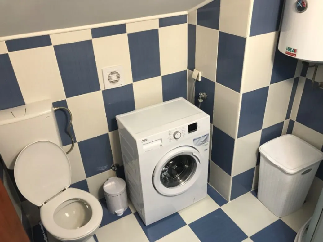 Kupatilo sa plavo-bijelim pločicama, wc šoljom, mašinom za veš i bojlerom.