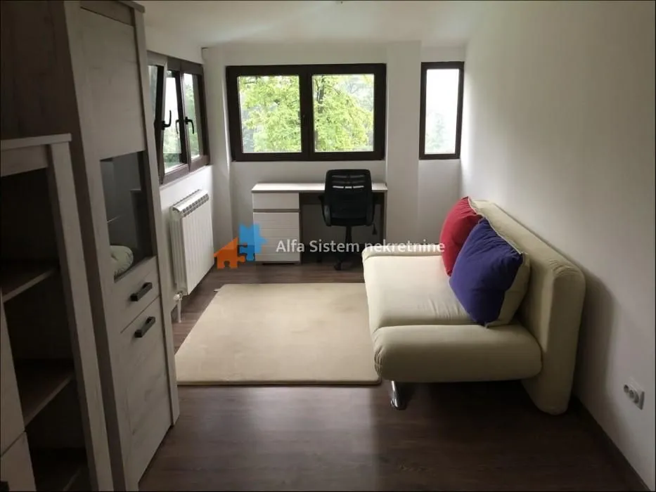 Living room with a sofa, desk, and windows overlooking greenery.