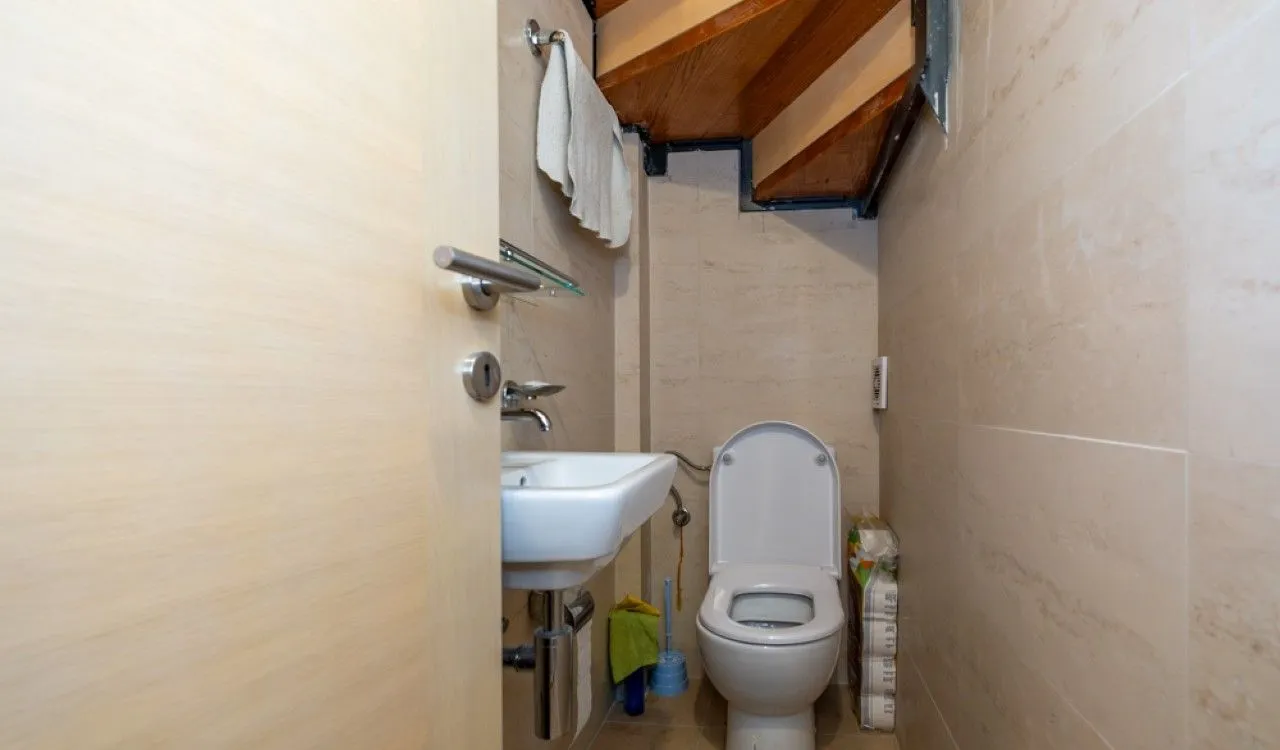 Manje kupatilo sa WC šoljom, umivaonikom i drvenim stepeništem iznad.