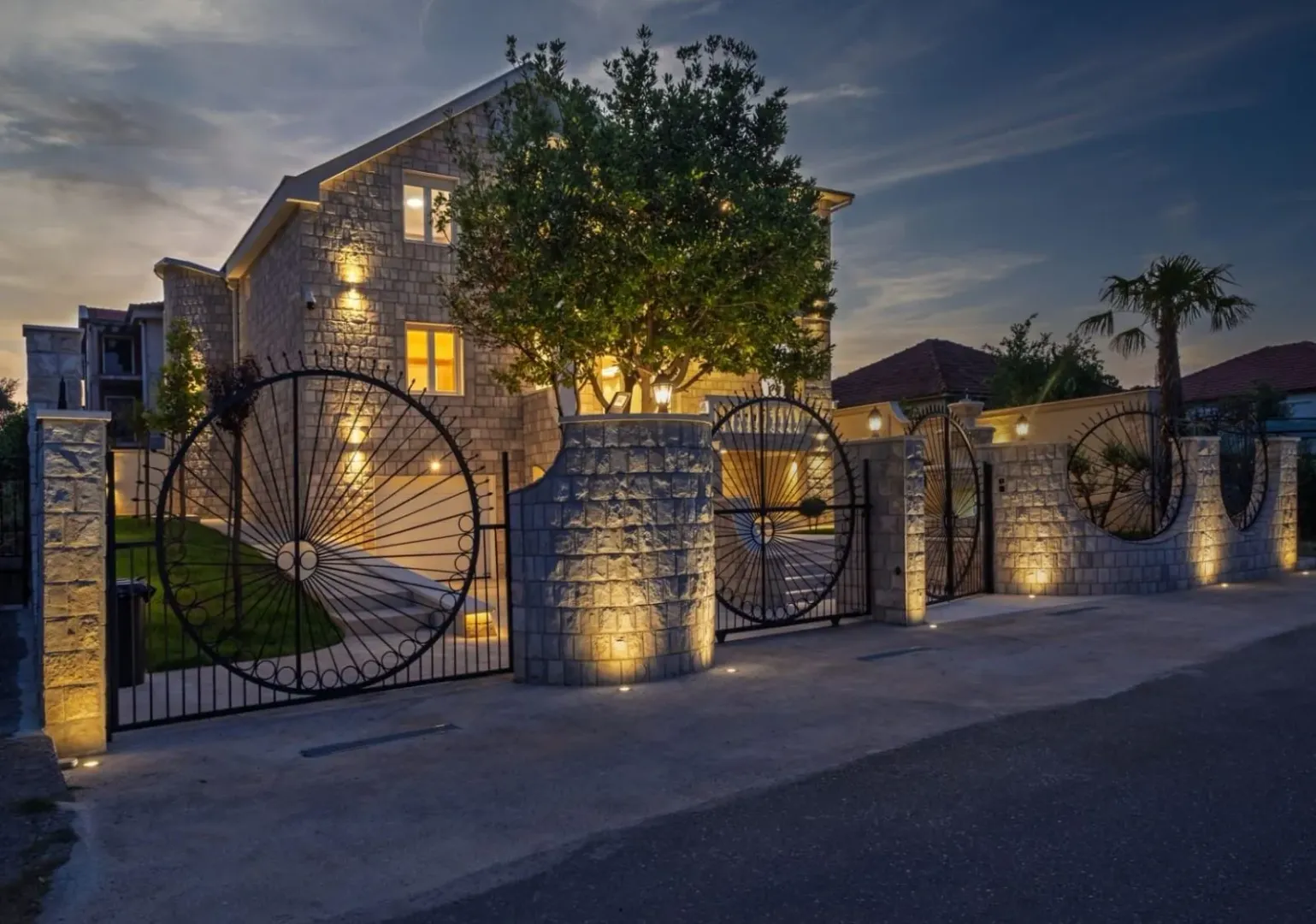 View of the illuminated house facade with courtyard and fence at twilight.