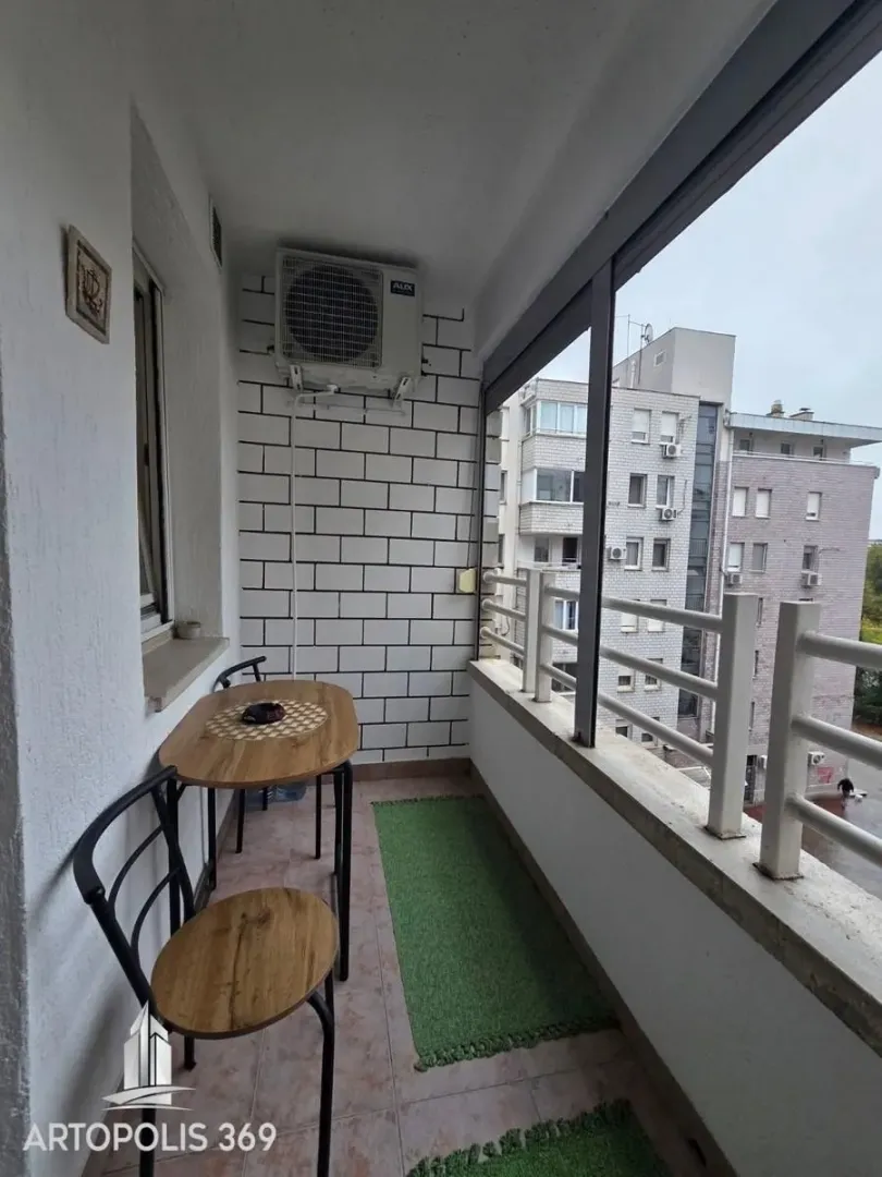 Natkriveni balkon sa garniturom za sedenje i pogledom na okolne zgrade.
